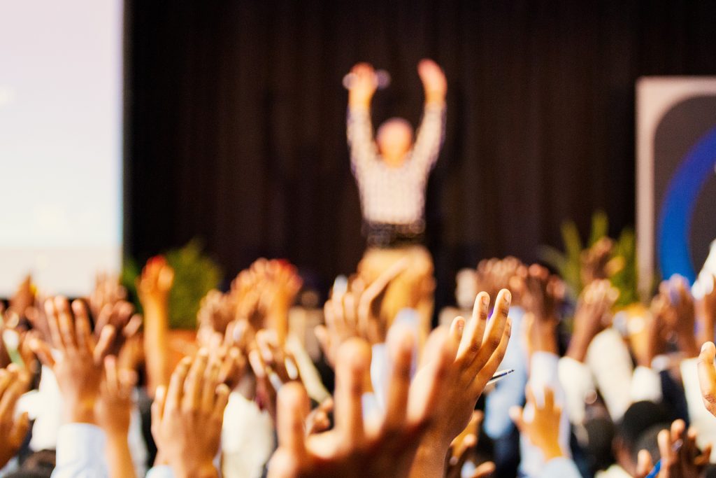 Artistic photo of a performer on stage with their hands up with the crowd with some hands in focus.