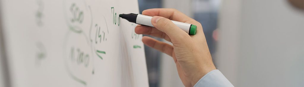 Photo of a hand holding a green dry erase marker, writing on a whiteboard