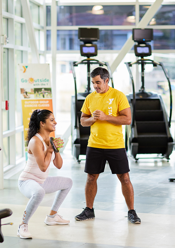 Mujer haciendo sentadillas mientras es entrenada por un entrenador de la YMCA en el gimnasio local.
